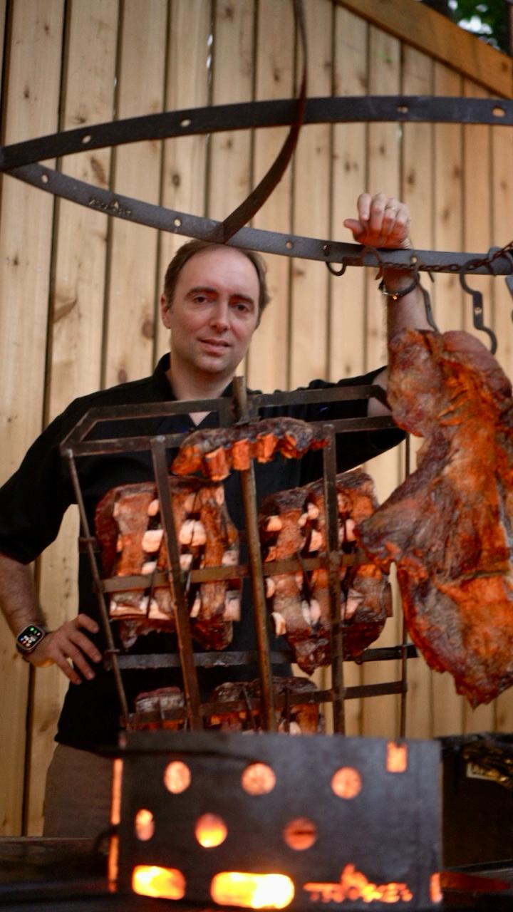 Man standing next to a large metal rack with hanging meat over an open fire.